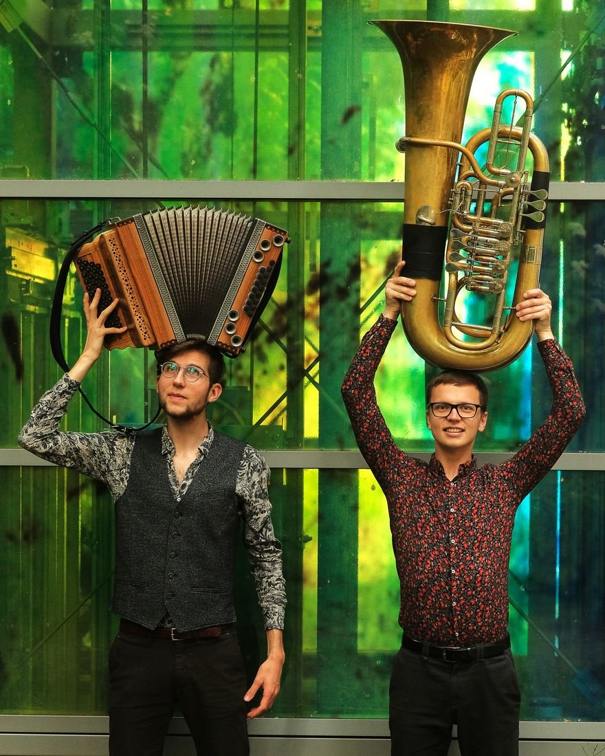 Jonny Kölbl hält eine Steirische Harmonika, Tobias Weiß eine Tuba über dem Kopf, vor einer grünen Glasfassade.